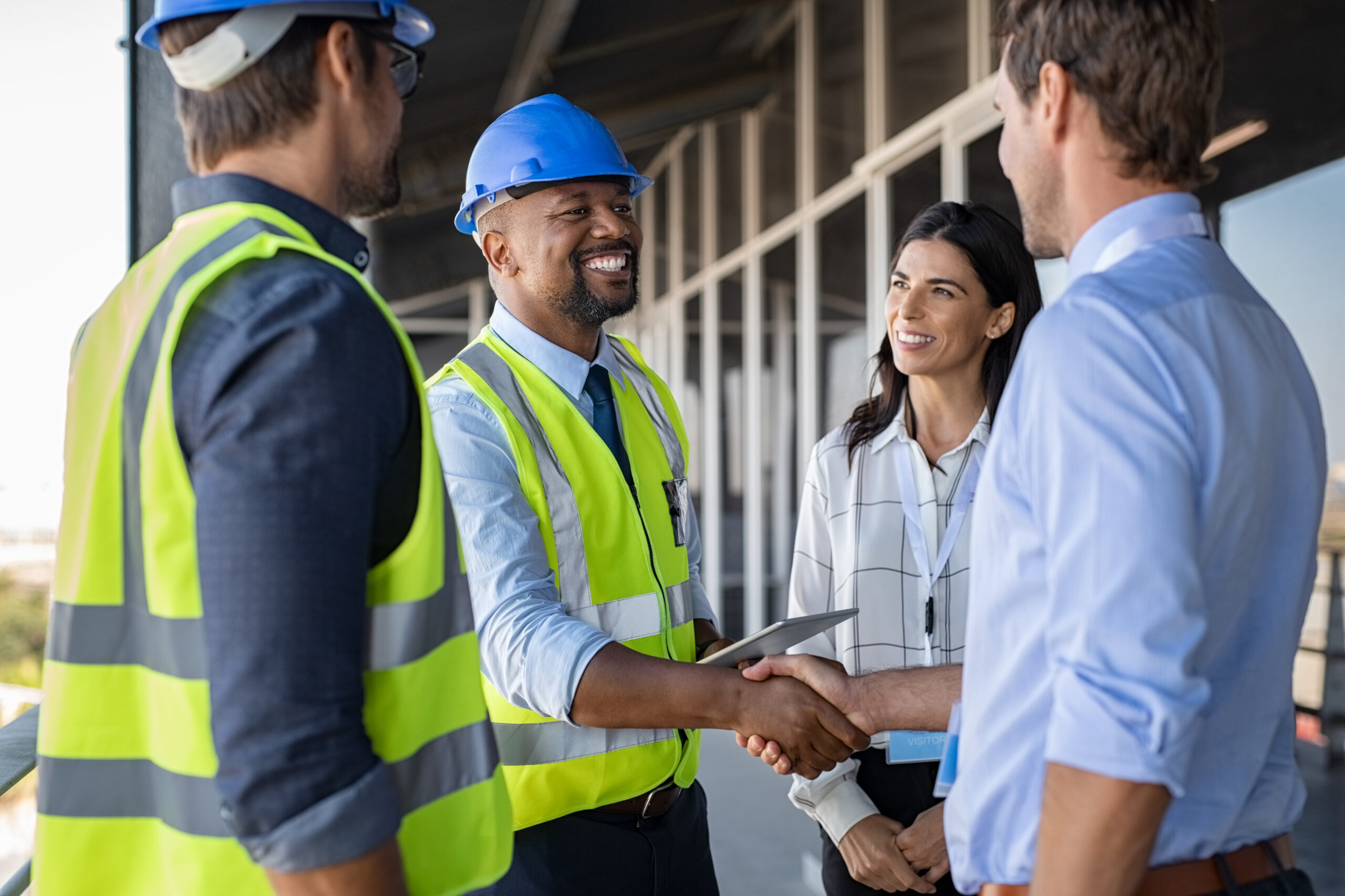 Construction team members engage in a handshake, discussing project details, while wearing safety gear. The setting is a construction site, emphasizing collaboration.