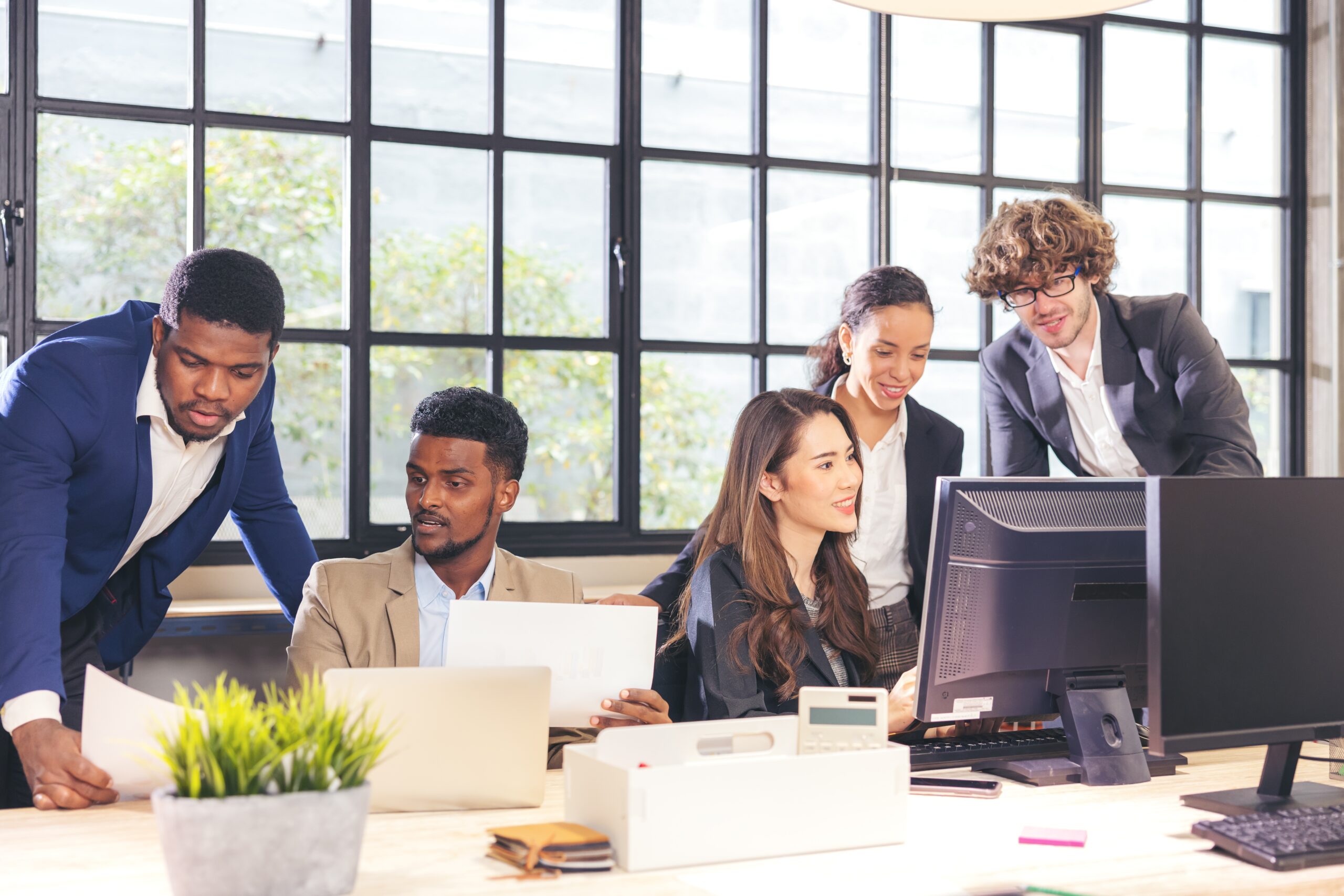 A diverse group of five professionals collaborates at a modern office, discussing work around a laptop and desktop computer.