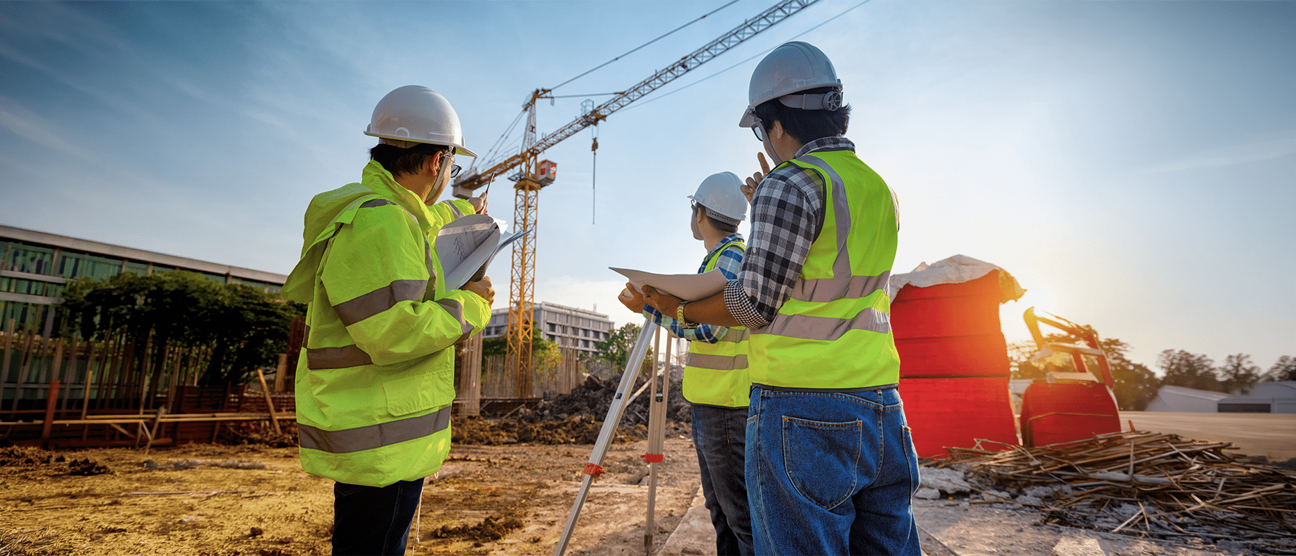 Three construction professionals in safety vests and helmets survey a building site, using plans and survey equipment as a crane operates nearby.