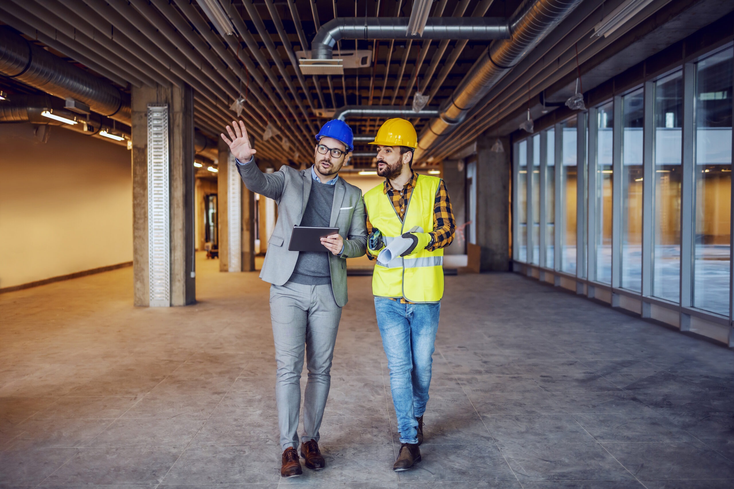 Two men converse in a partially finished construction site. One, in a suit, holds a tablet; the other, in workwear, gestures while discussing plans.