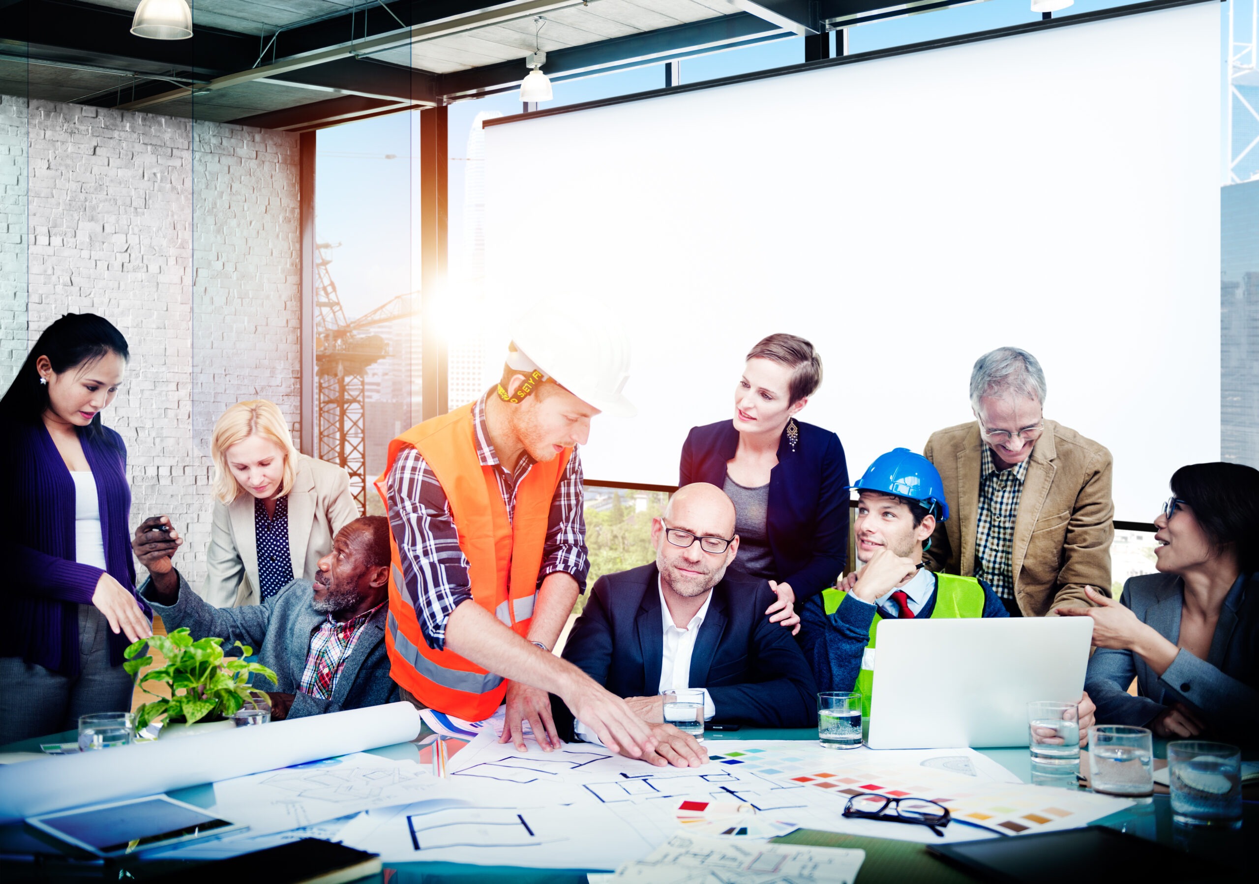 A diverse group of professionals gather around a table with blueprints, discussing a construction project. A worker in a hard hat and safety vest points at plans, engaging others.
